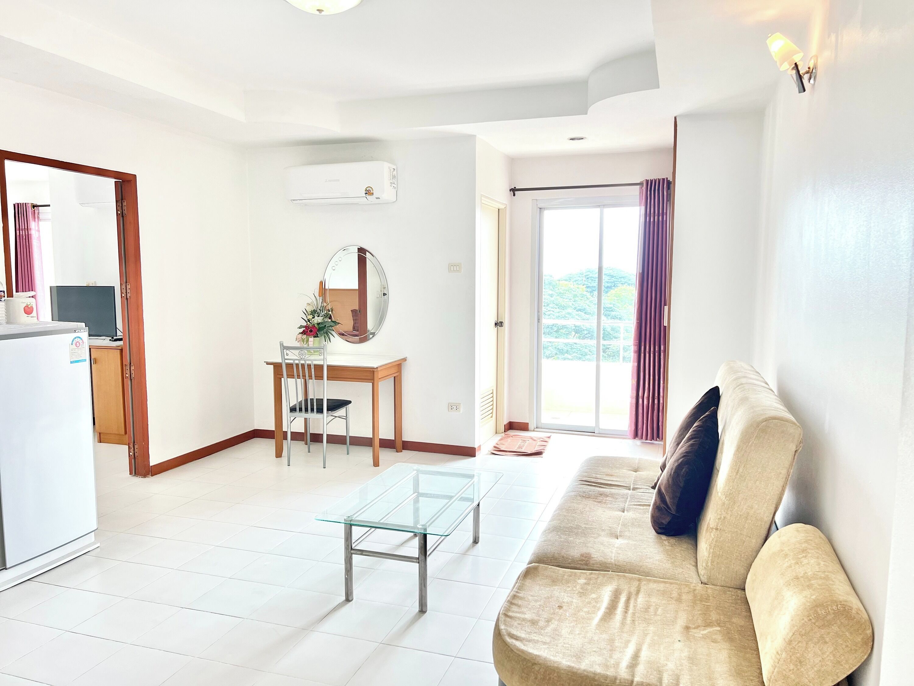 A living room with a sofa, a glass coffee table, a desk with a chair, a mirror, and a sliding glass door leading to a balcony.