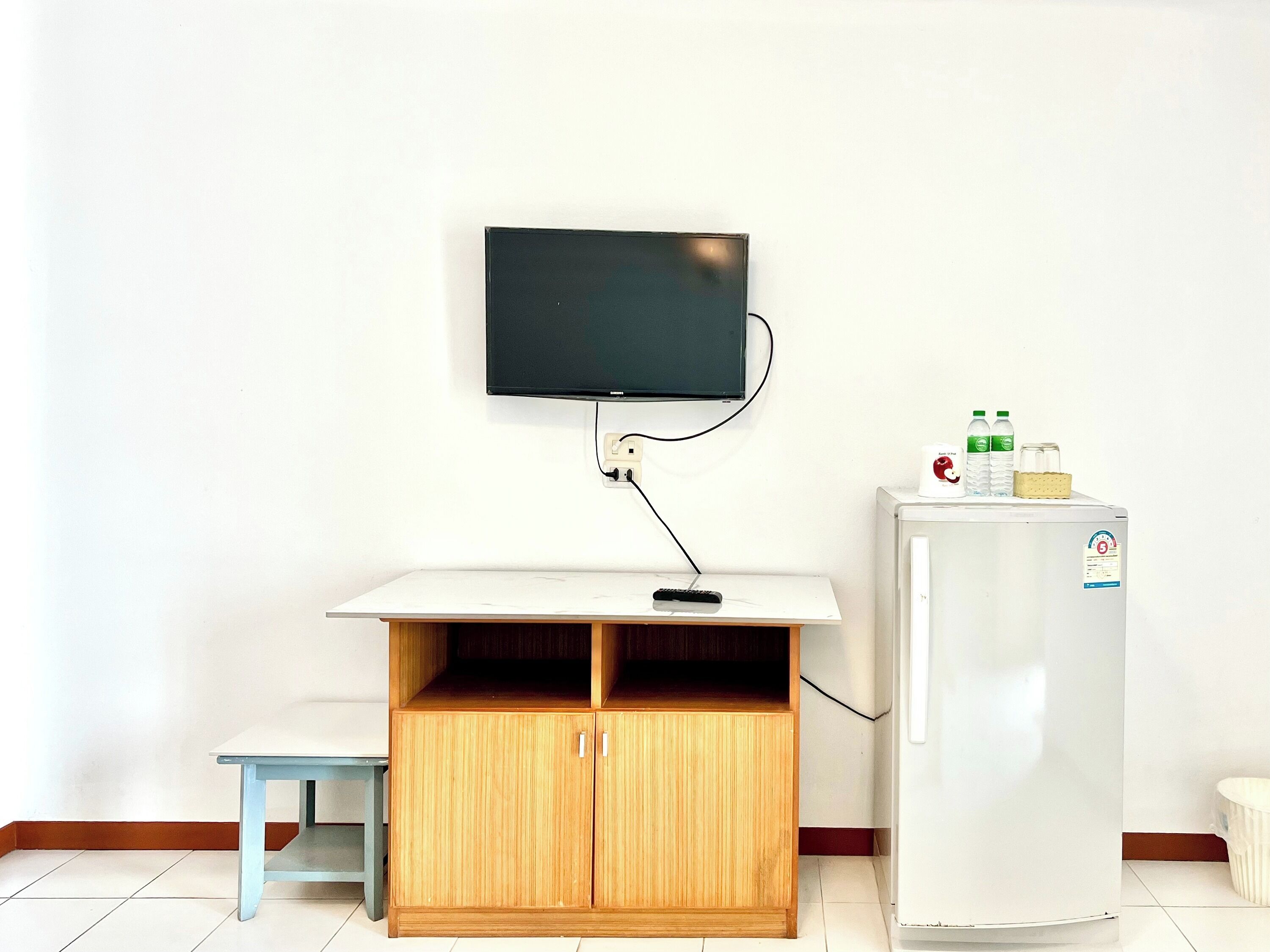 A room with a television mounted on the wall, a wooden cabinet with two doors, a small table, and a refrigerator.