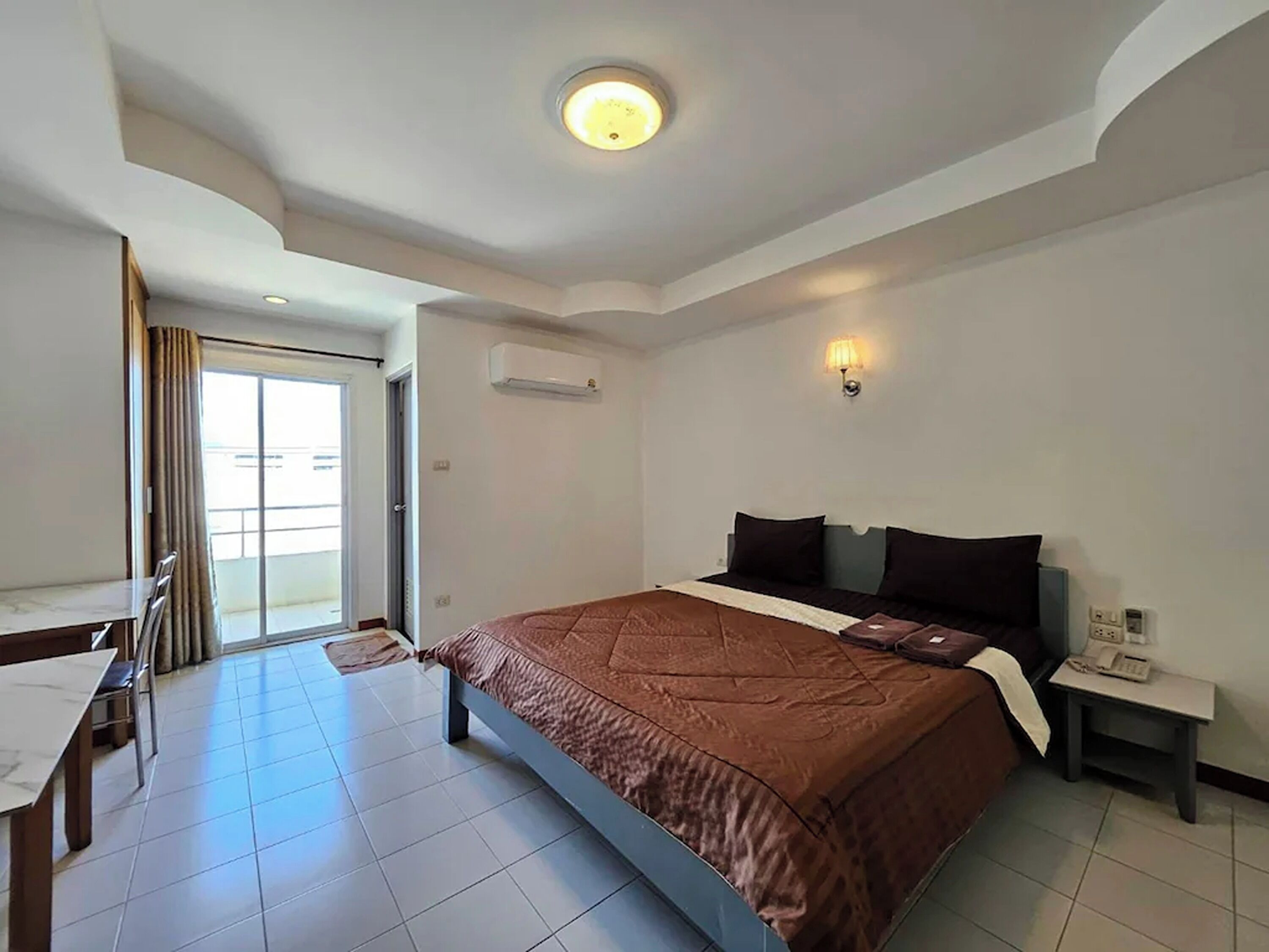 Habitación de hotel con una cama grande, un escritorio con silla, una mesita, una mesita de noche y una lámpara de pared.