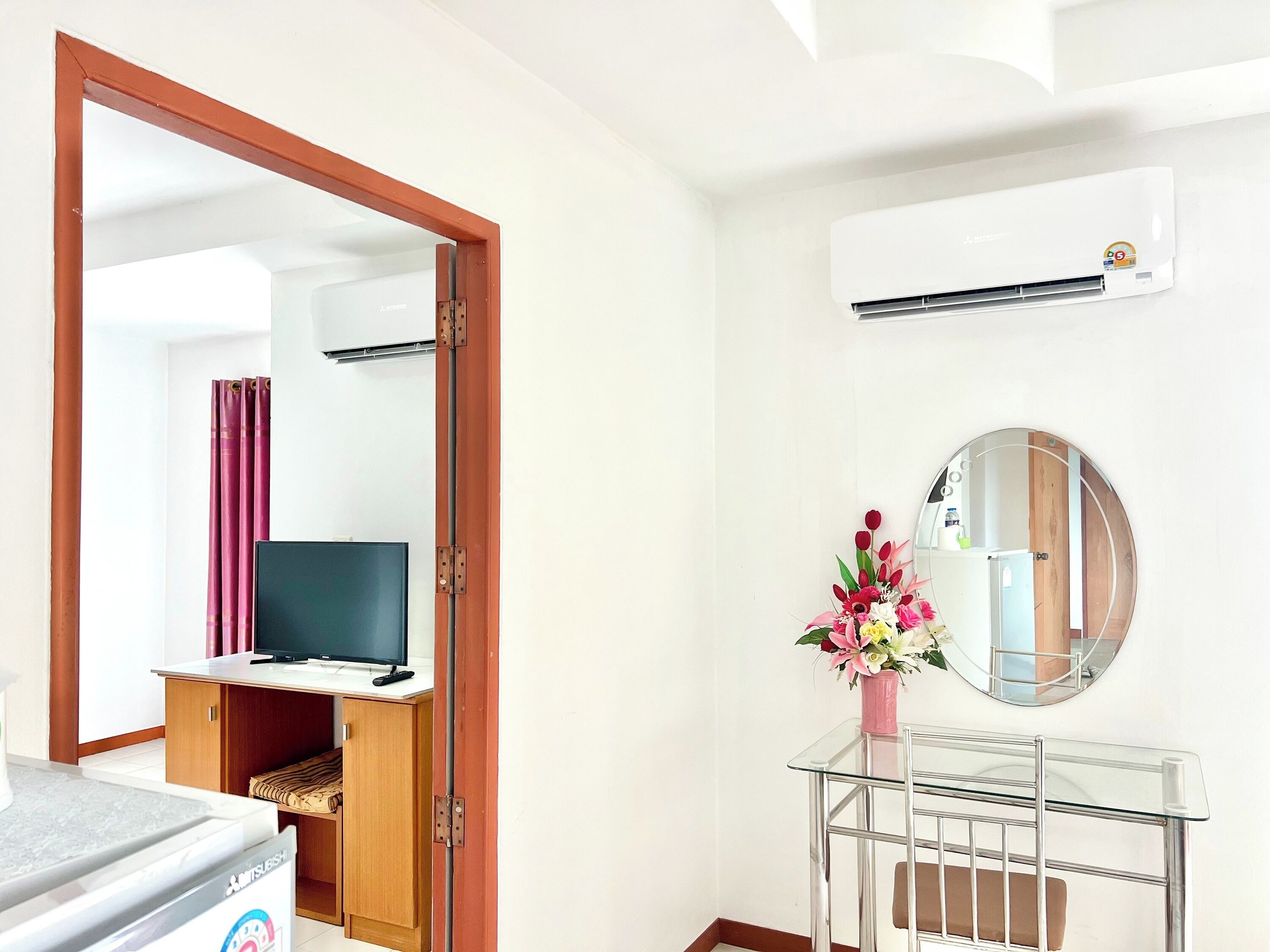 A hotel room with a bed, a TV, a desk with a computer, a mirror, and an air conditioning unit.
