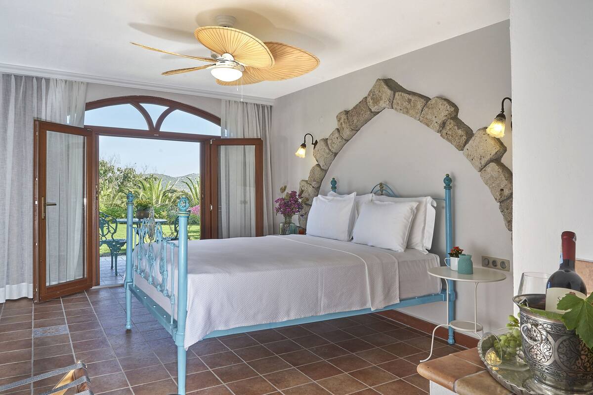 Een slaapkamer met een bed, een plafondventilator, een muur van natuursteen, een klein tafeltje met een lamp, en uitzicht op de omgeving.