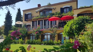 A two-story building with a yellow facade, multiple balconies with red umbrellas, and a well-maintained garden with various plants and flowers. - Villa Taraca Alacati Romantik Hotel (Cesme)