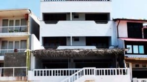 Un hotel frente a la playa con fachada blanca, un balcón y una escalera que baja a la arena. Hay sillas de playa azules sobre la arena.