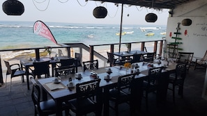 Een restaurant aan zee met terras en uitzicht op de oceaan, met een naambordje en zicht op boten in het water.