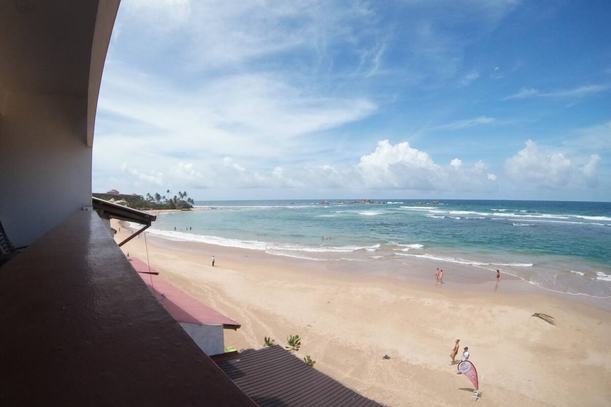 Een balkon met uitzicht op een strand, de oceaan en verre palmbomen.