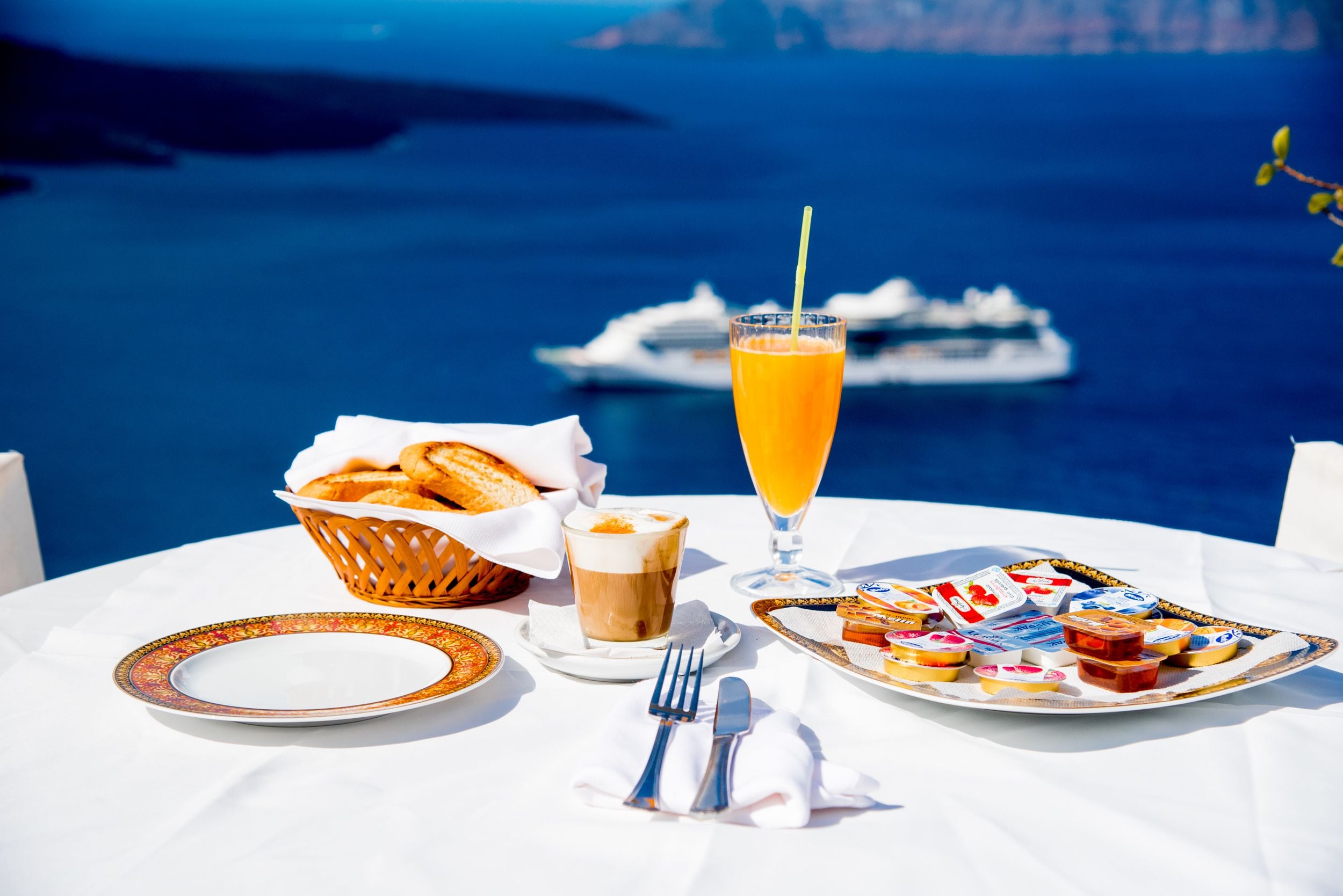 Una mesa preparada para el desayuno, un vaso de jugo de naranja y vista al mar con un crucero en el fondo.