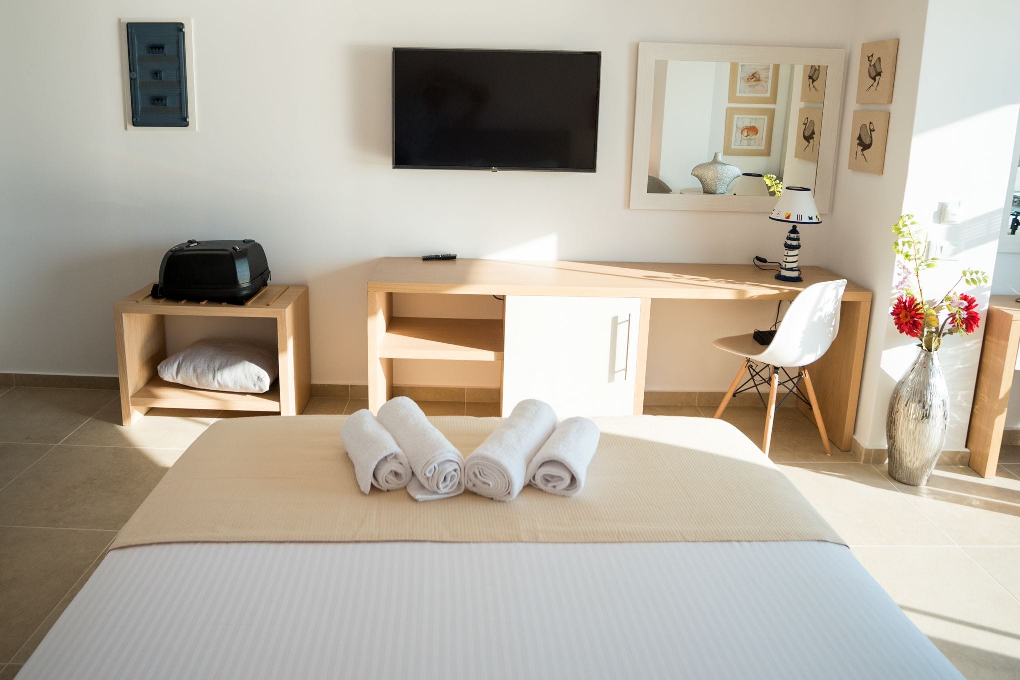 Habitación de hotel con cama, escritorio, televisor, lámpara y un florero con flores.