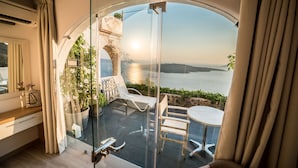 A balcony with a glass door, a view of the sea, and a small table with chairs. - Kratiras View Luxury Suites (Santorini)
