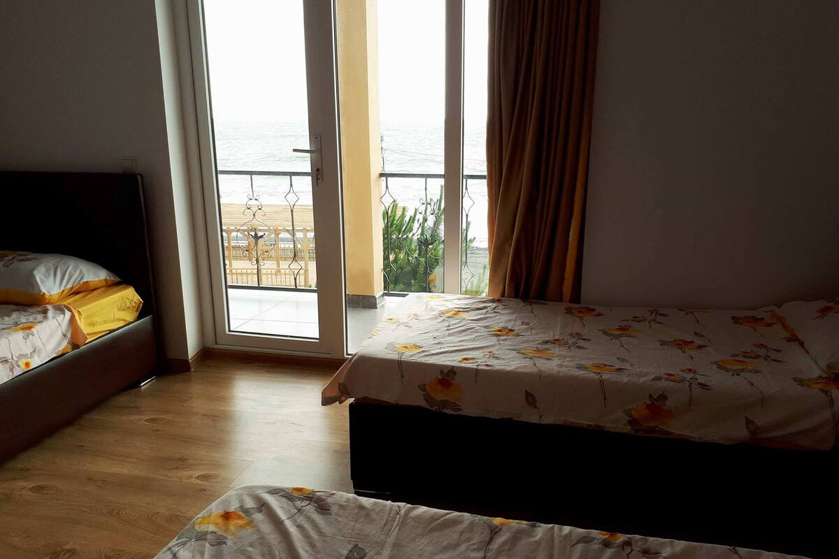 A room with two beds, a balcony with a view of the sea, and a sliding glass door.