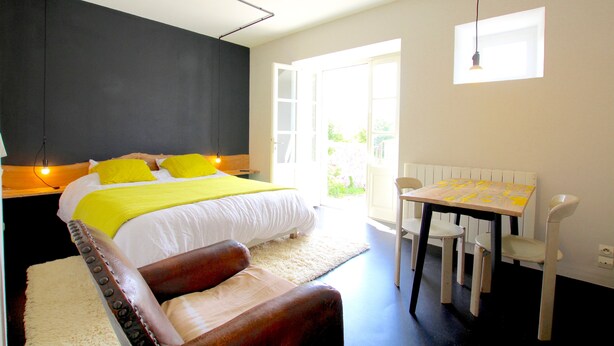 A bedroom with a bed, bedside table, and a small table with chairs. There is a window providing natural light. - La Maison Folia (Quettreville-sur-Sienne)