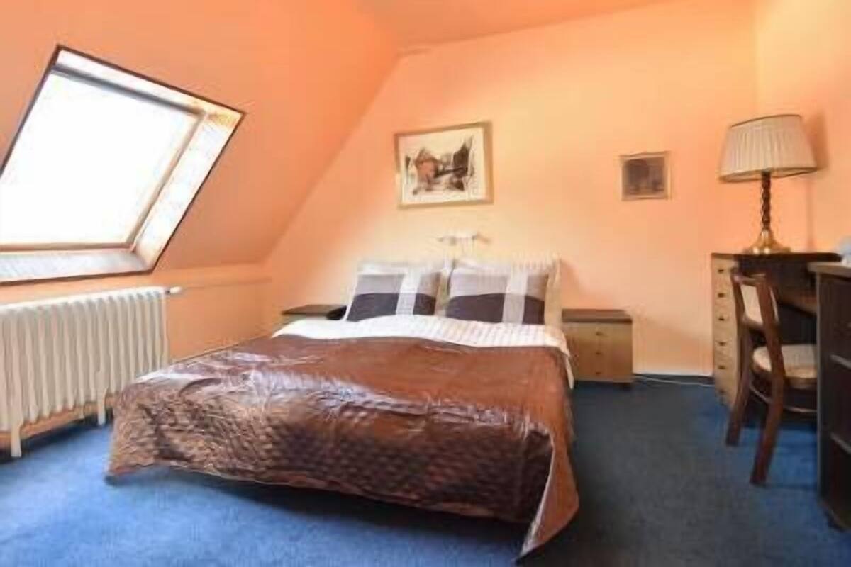 A bedroom with a bed, a desk, a chair, a radiator, a window, and a picture on the wall.