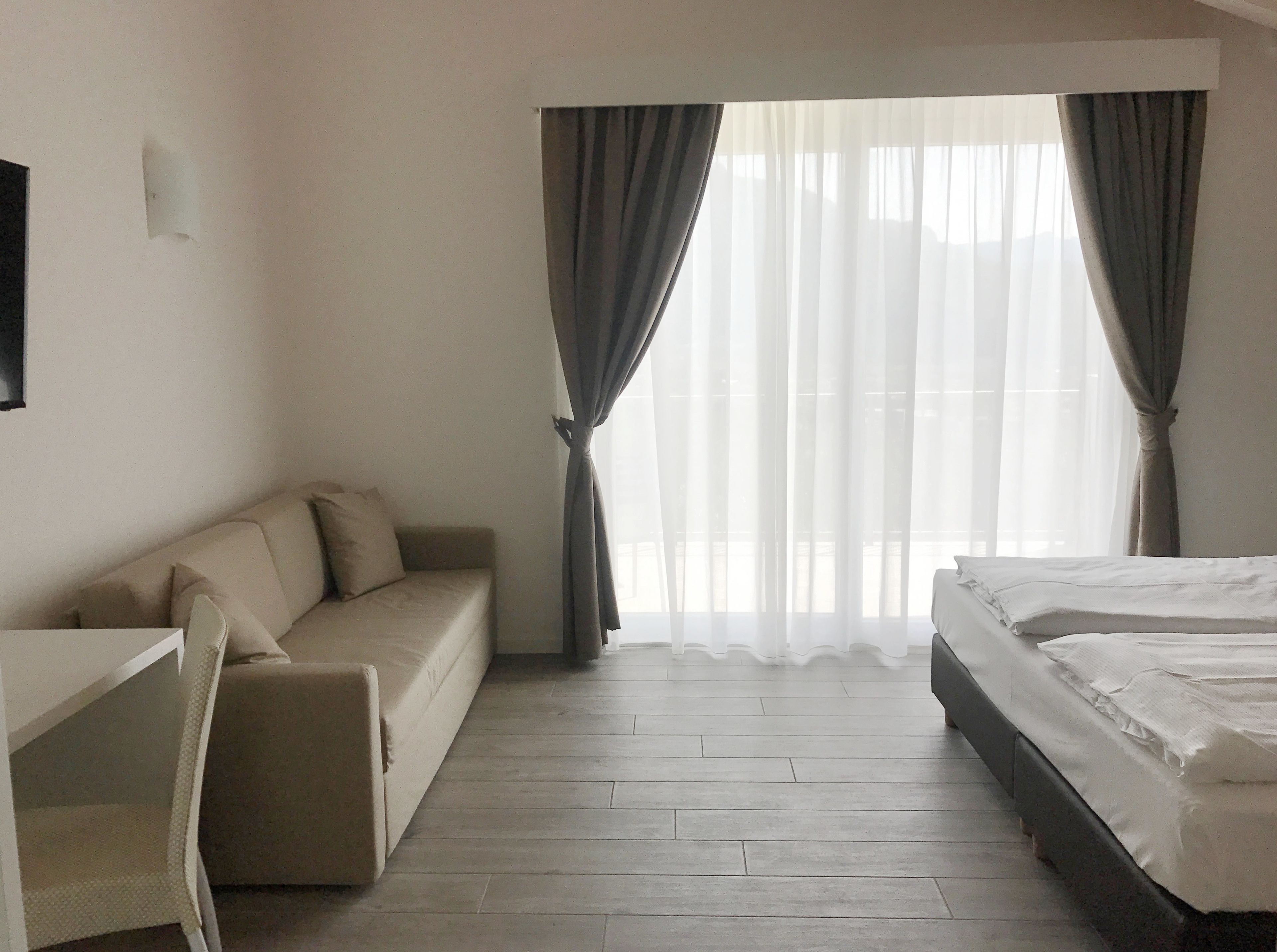 A hotel room with a bed, a sofa, a desk, and a large window with curtains.