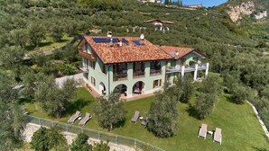 Une maison à deux étages, dotée d’un toit en tuiles, entourée d’une végétation luxuriante et de collines.