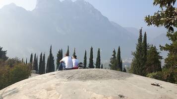 A large concrete viewing platform with two people sitting, overlooking a mountainous landscape with tall trees and a distant city.