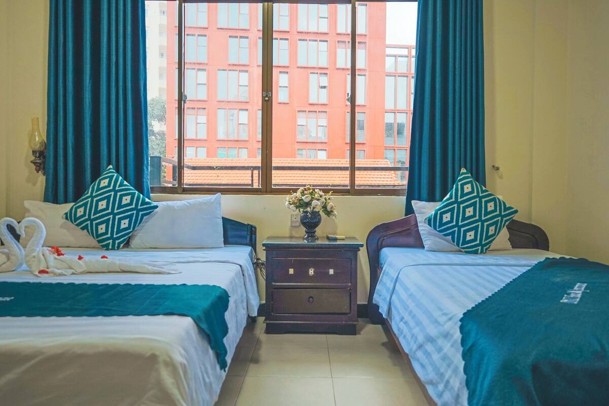 Habitación de hotel con dos camas, una mesita de noche con un florero, y vista a un edificio rojo a través de la ventana.