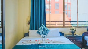 Habitación de hotel con una cama, una mesita de noche con un jarrón de flores y una ventana con vistas a edificios.
