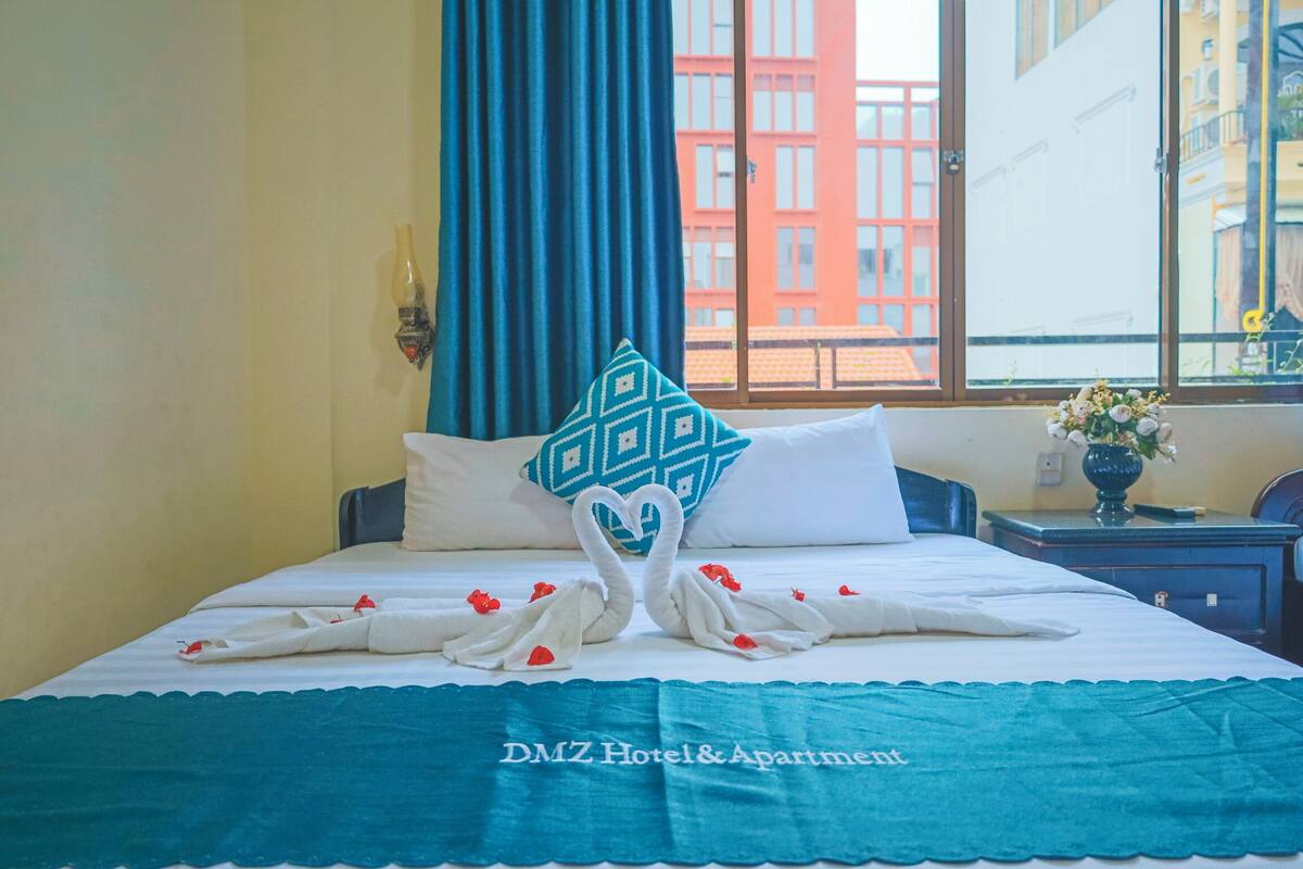 Habitación de hotel con cama, cortina azul, almohada decorativa y un florero con flores sobre una mesita de noche.