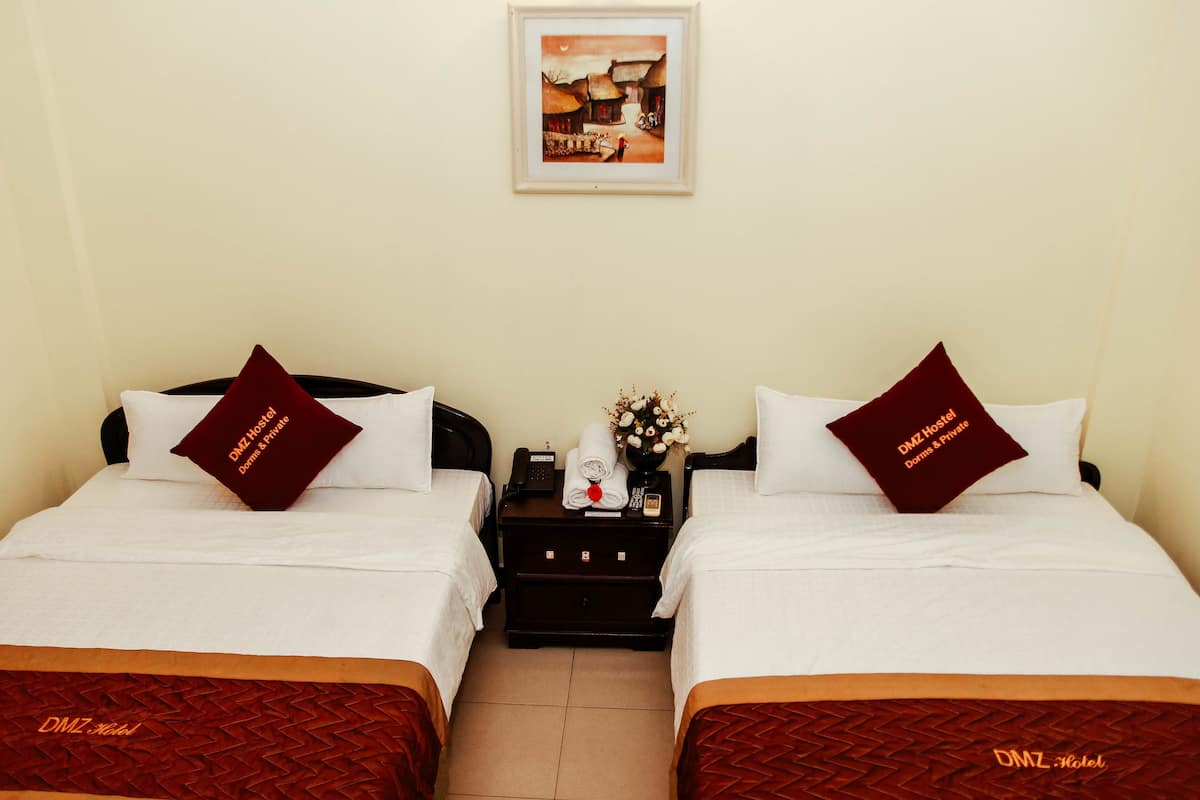 Habitación de hotel con dos camas, cada una con una almohada que dice "DMZ Hotel". Un mesita de noche con un teléfono y un florero se encuentra entre las camas.