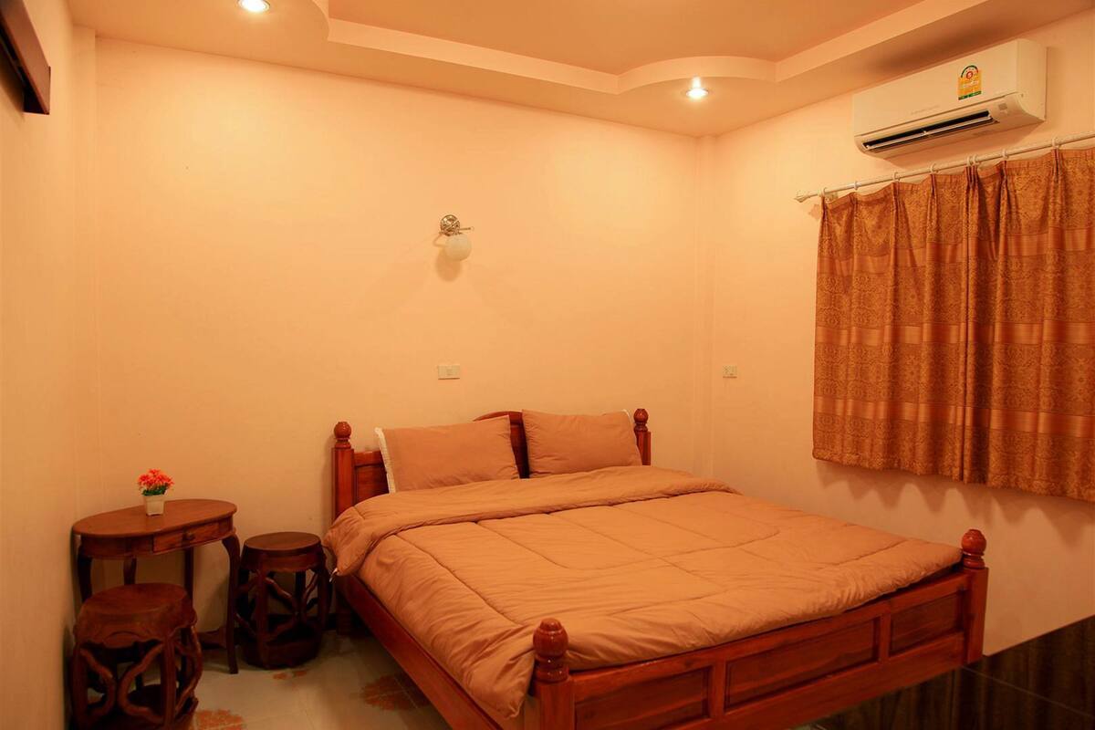 A bedroom with a wooden bed, a small round table with a flower, two stools, a wall-mounted air conditioner, and a curtain.