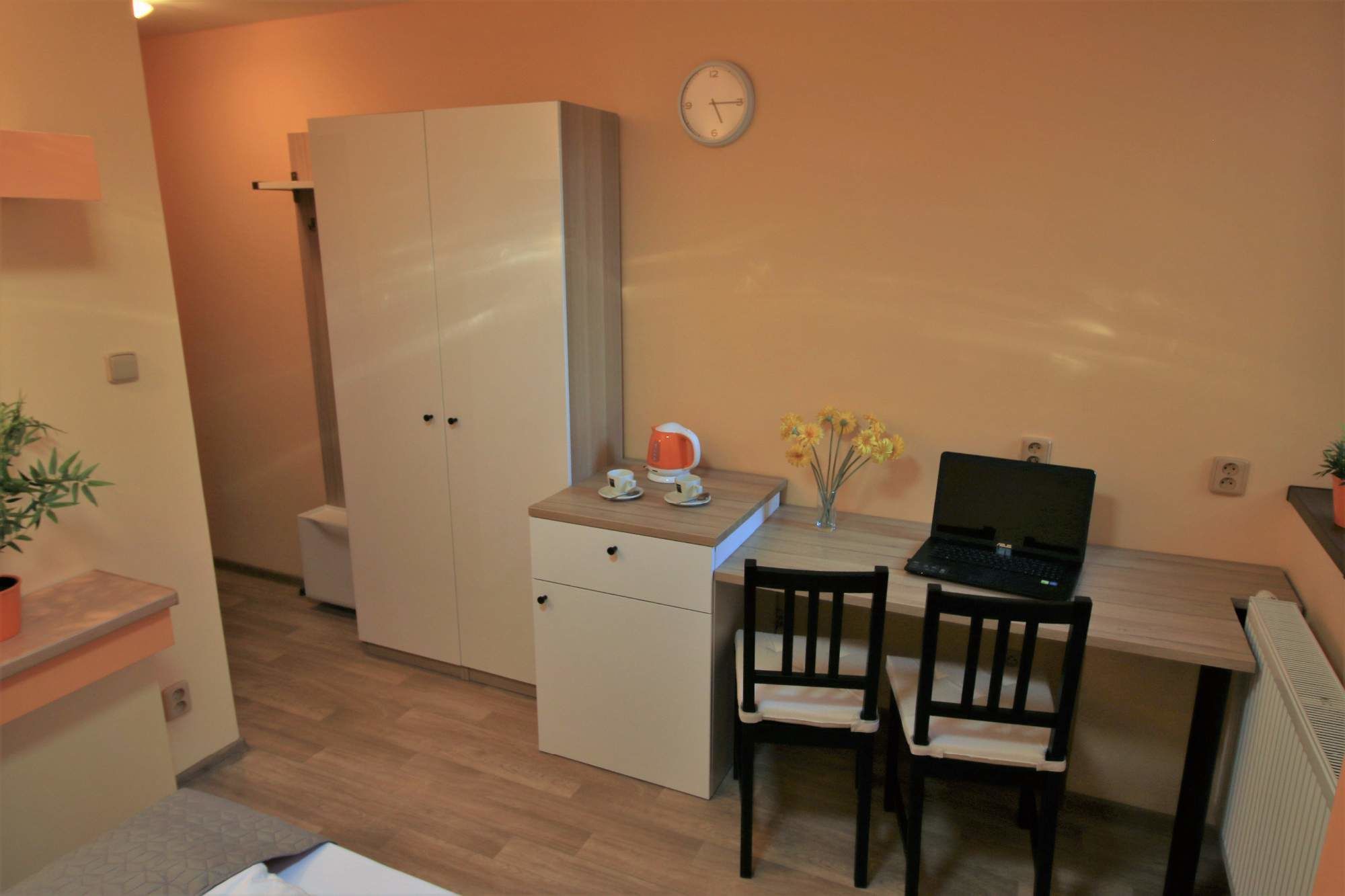 A small, compact room with a kitchenette, a desk with two chairs, a clock on the wall, and a radiator.