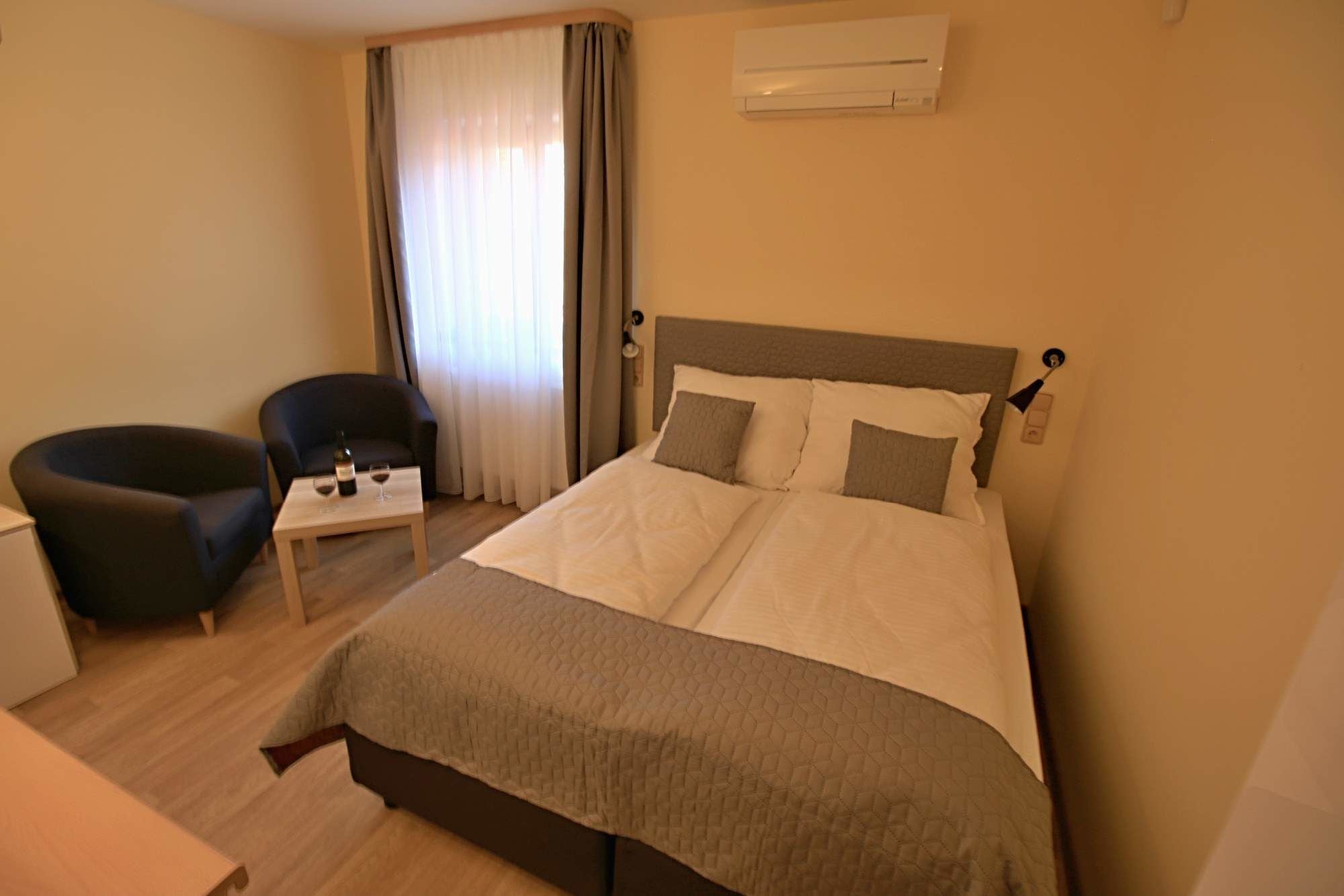 A hotel room with a bed, a chair, a small table with a bottle and glass, and an air conditioning unit on the wall.