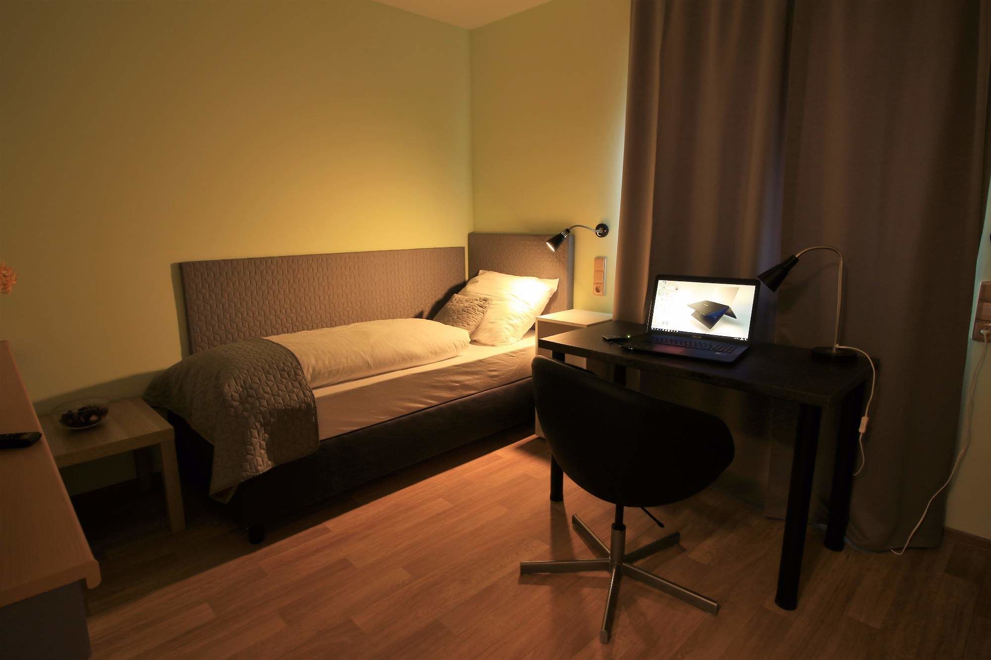 A small, well-lit room with a bed, a desk with a laptop, and a chair.
