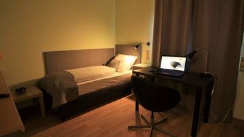 A small, well-lit room with a bed, a desk with a laptop, and a chair.