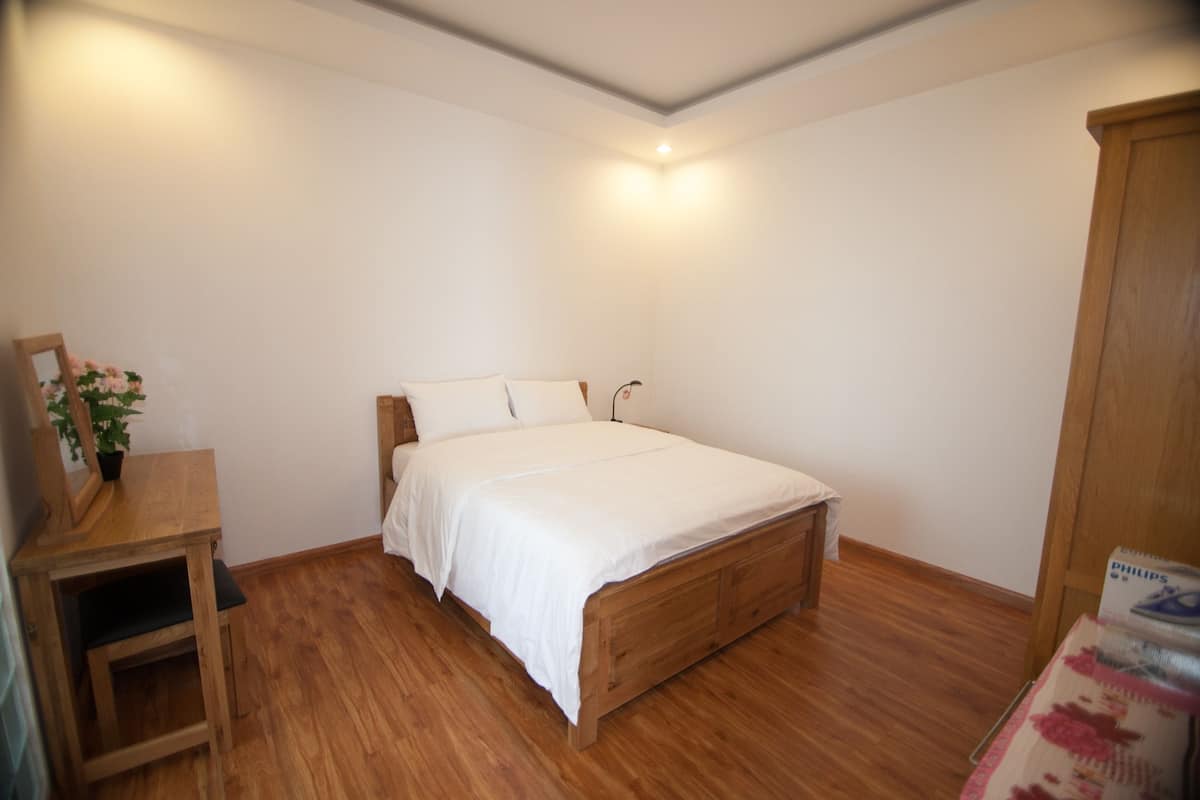 A bedroom with a wooden bed, white bedding, a small wooden table with a plant, and a wooden wardrobe.