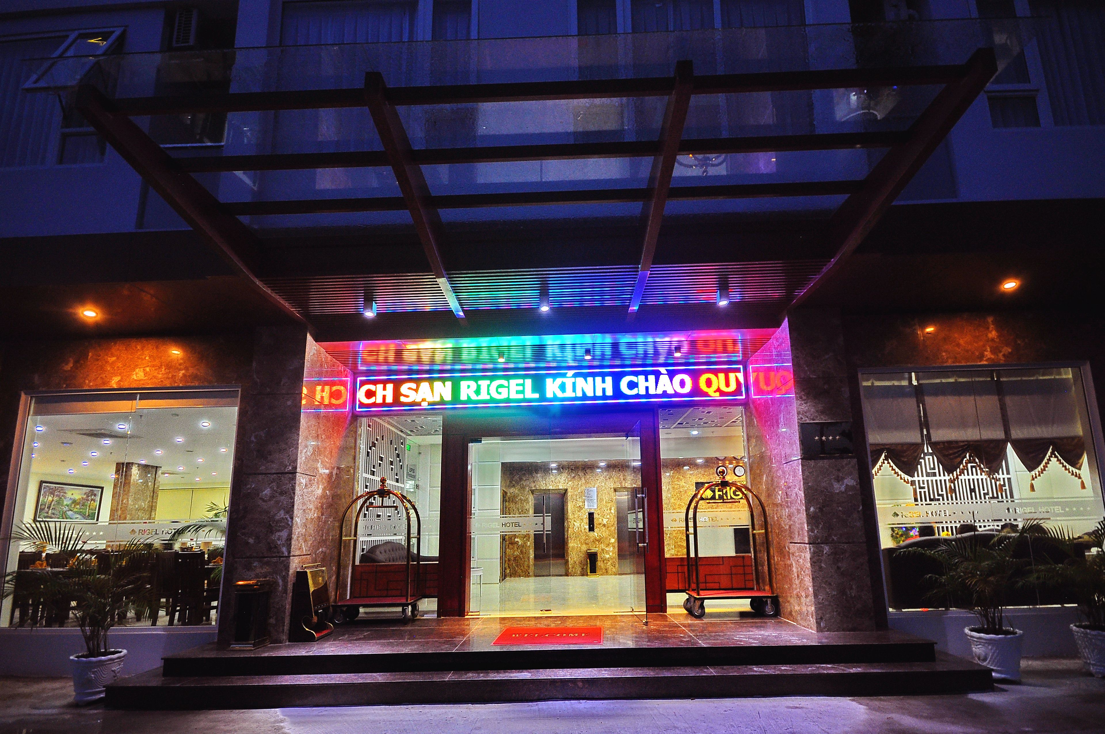 The entrance of a hotel with a neon sign, glass doors, and a ramp leading up to the entrance.