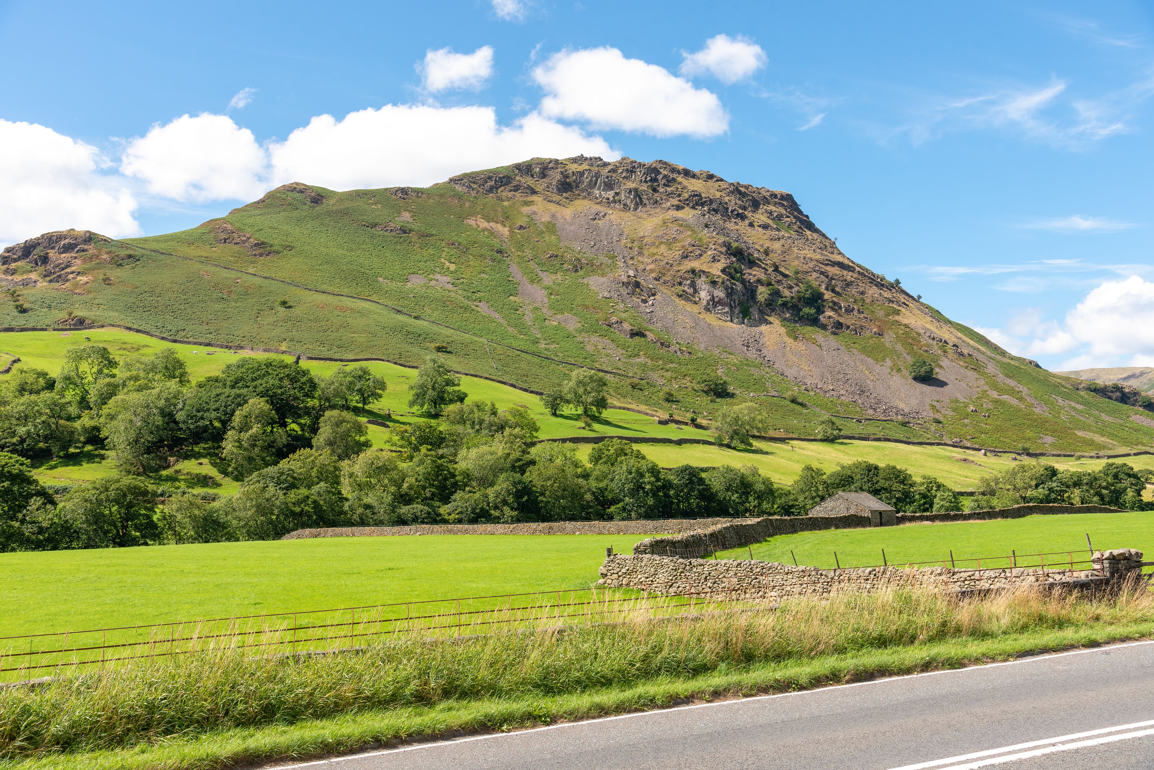 A rural landscape with a stone wall, a road, and a hill with trees.