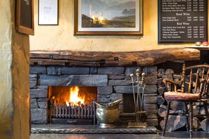 A stone fireplace with a lit fire, a metal bucket, and a wooden chair.