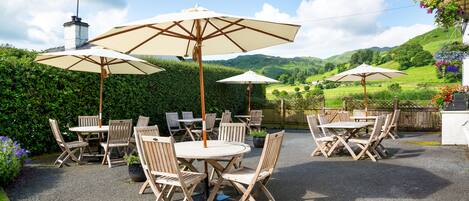 Une terrasse extérieure avec des tables et des chaises rondes, entourée de parasols, le tout se fondant dans un paysage de collines verdoyantes et d’un ciel dégagé.