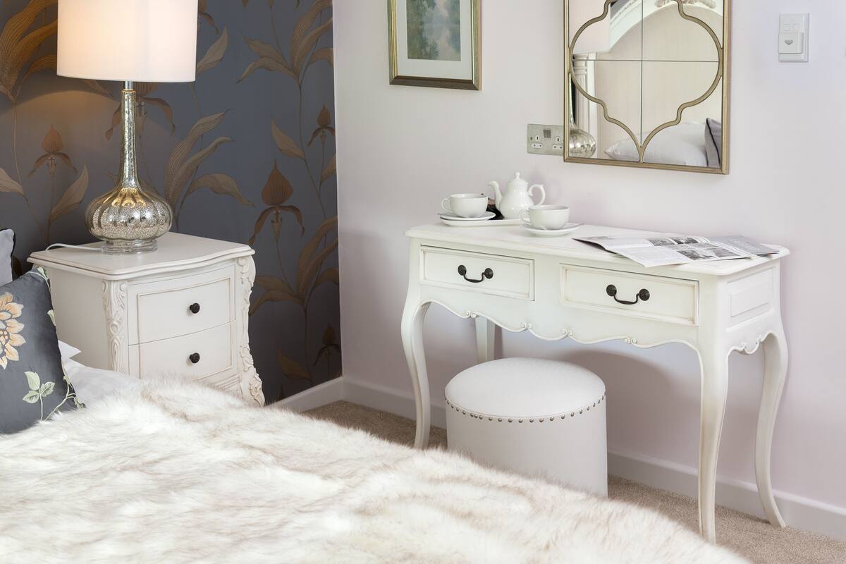A bedroom with a white vanity, a fluffy white bed, a lamp, and a mirror.
