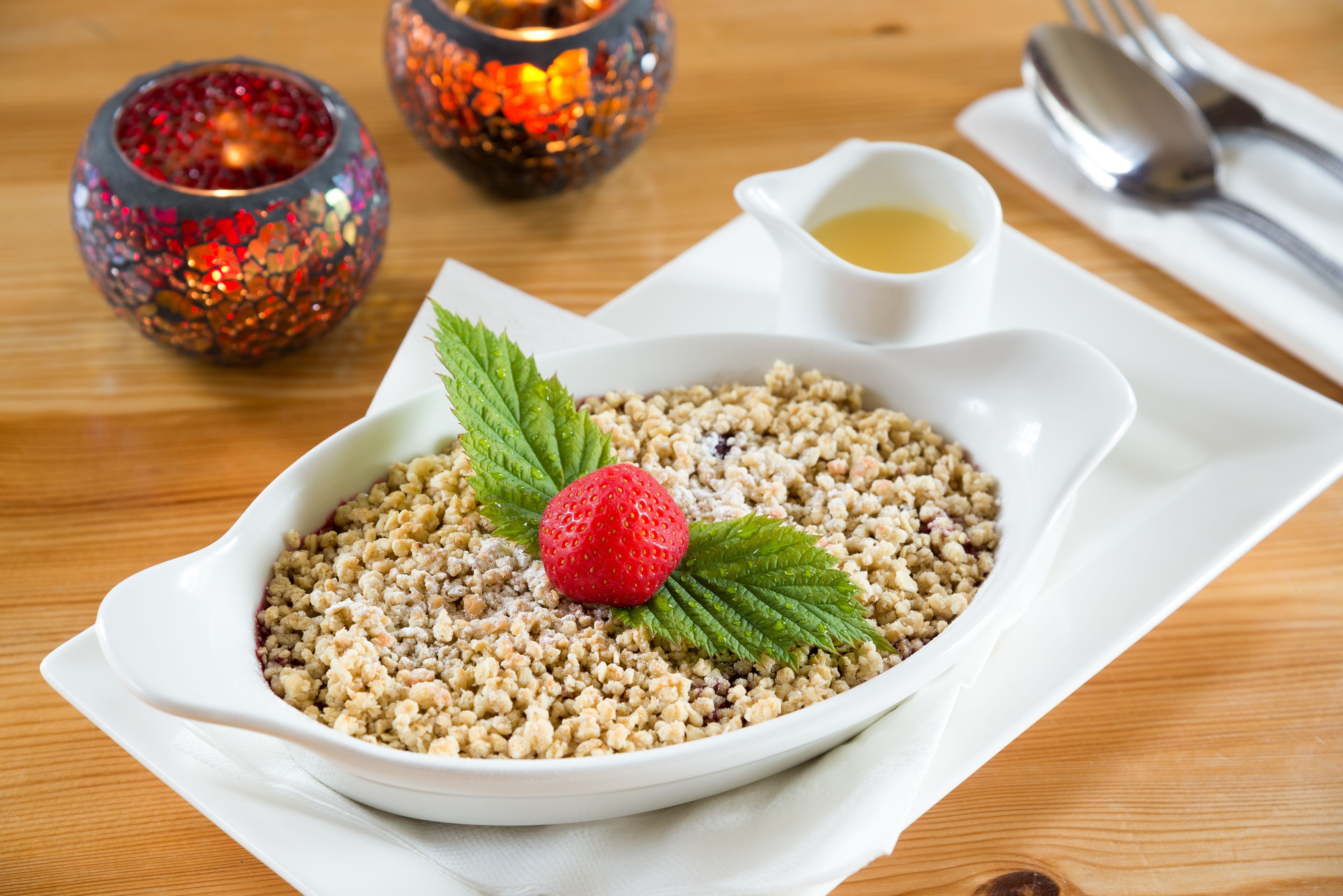 A white bowl of couscous garnished with a strawberry and mint leaves, accompanied by a small bowl of sauce, on a wooden table with a candle and cutlery.