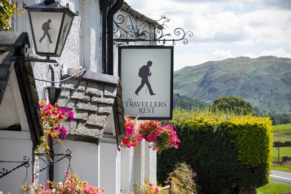 A sign for 'The Travellers Rest' with a walking figure, surrounded by flowers and a mountainous backdrop.