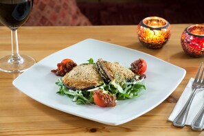 A square white plate with a sandwich and salad on a wooden table, accompanied by a wine glass and two decorative candles.