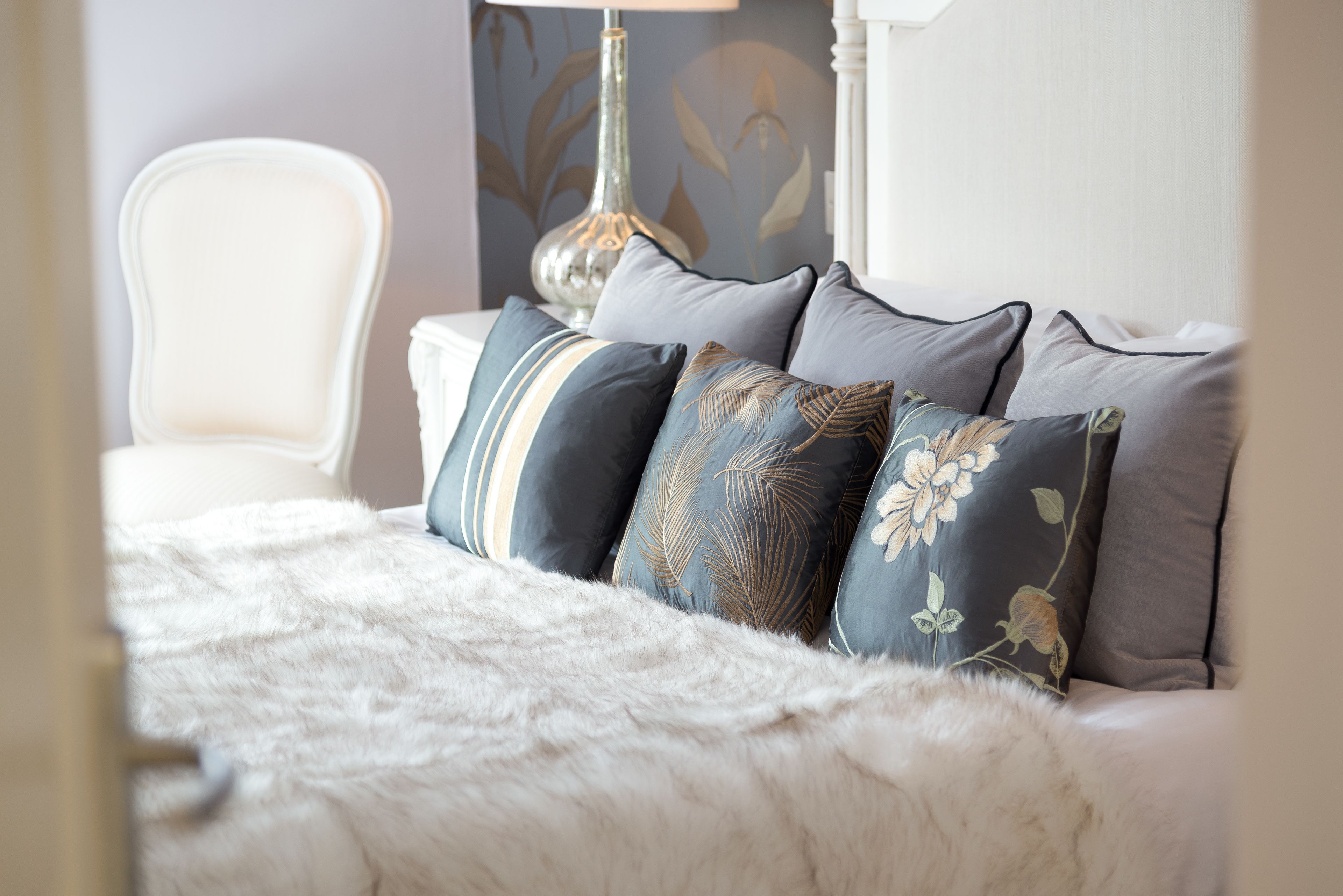 A bed with a white furry blanket, a white chair, and a lamp with a floral design on the wall behind.