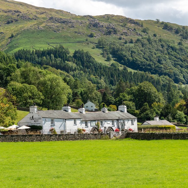 A white building with a sign reading "The Travellers Rest" in a rural setting with green fields and a forested hill in the background.