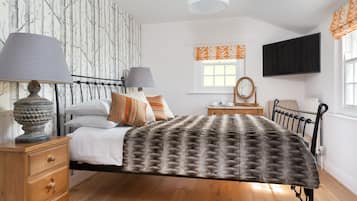 A bedroom with a bed, two bedside tables with lamps, a dresser, a TV mounted on the wall, a window with curtains, and a wall with a tree pattern wallpaper.