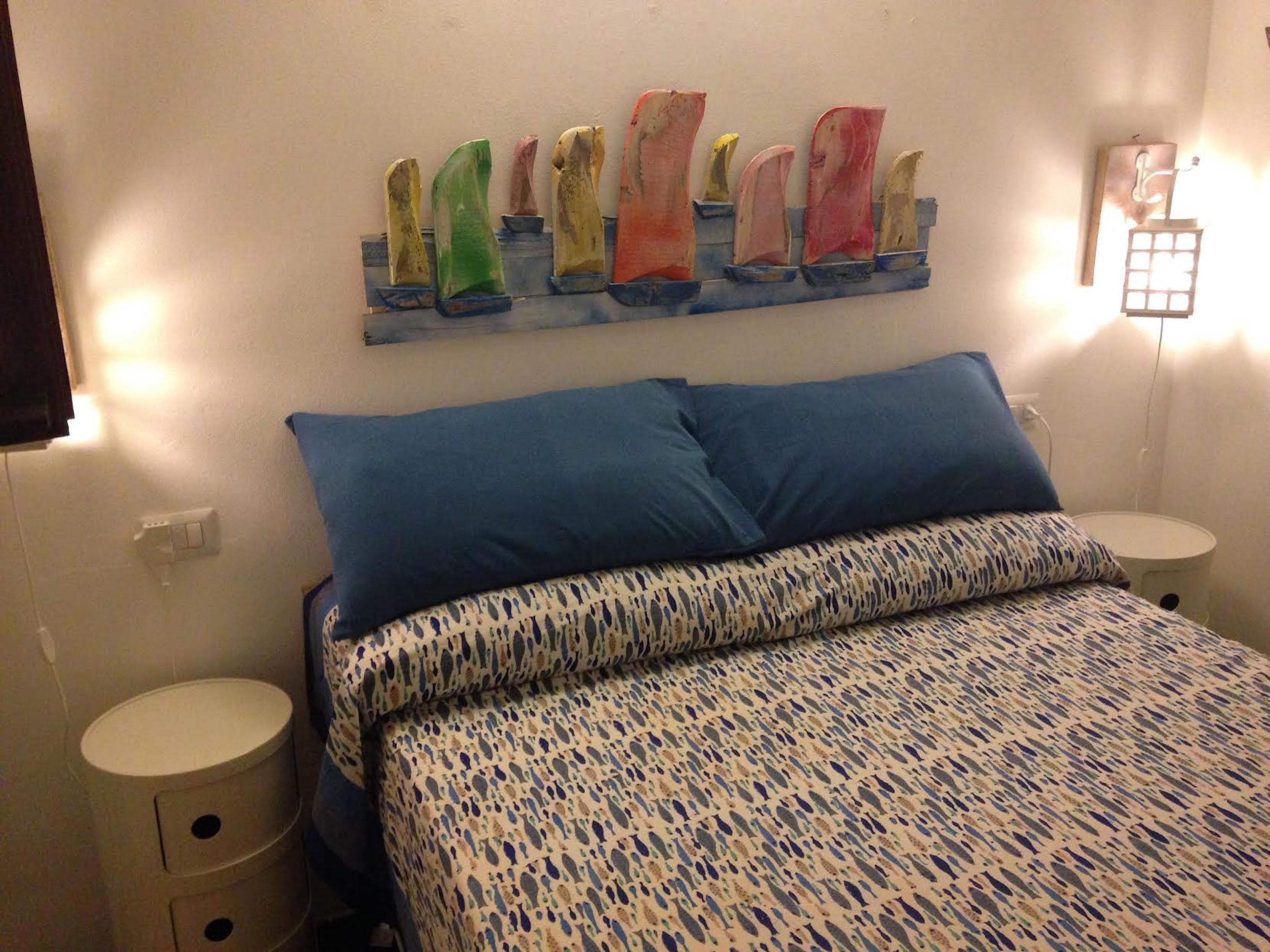 A bedroom with a bed, two nightstands, a wall-mounted light fixture, and a decorative wall piece.