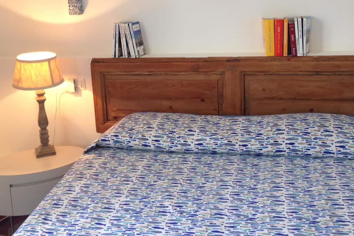 A bedroom with a wooden headboard, a patterned bedspread, a wall-mounted woven decoration, a bedside lamp, and a bookshelf with books.
