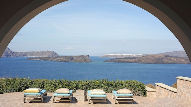 Une vue à travers une fenêtre en arc donnant sur un paysage côtier, avec trois chaises longues et un mur de pierre.