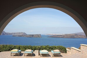 Une vue à travers une fenêtre en arc donnant sur un paysage côtier, avec trois chaises longues et un mur de pierre.