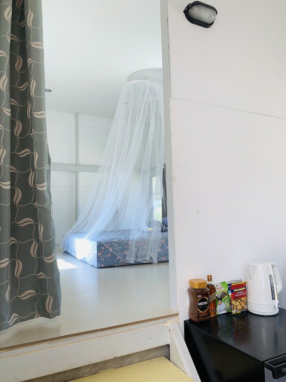 A room with a bed under a mosquito net, a window, and a kitchenette with a coffee maker and food items.