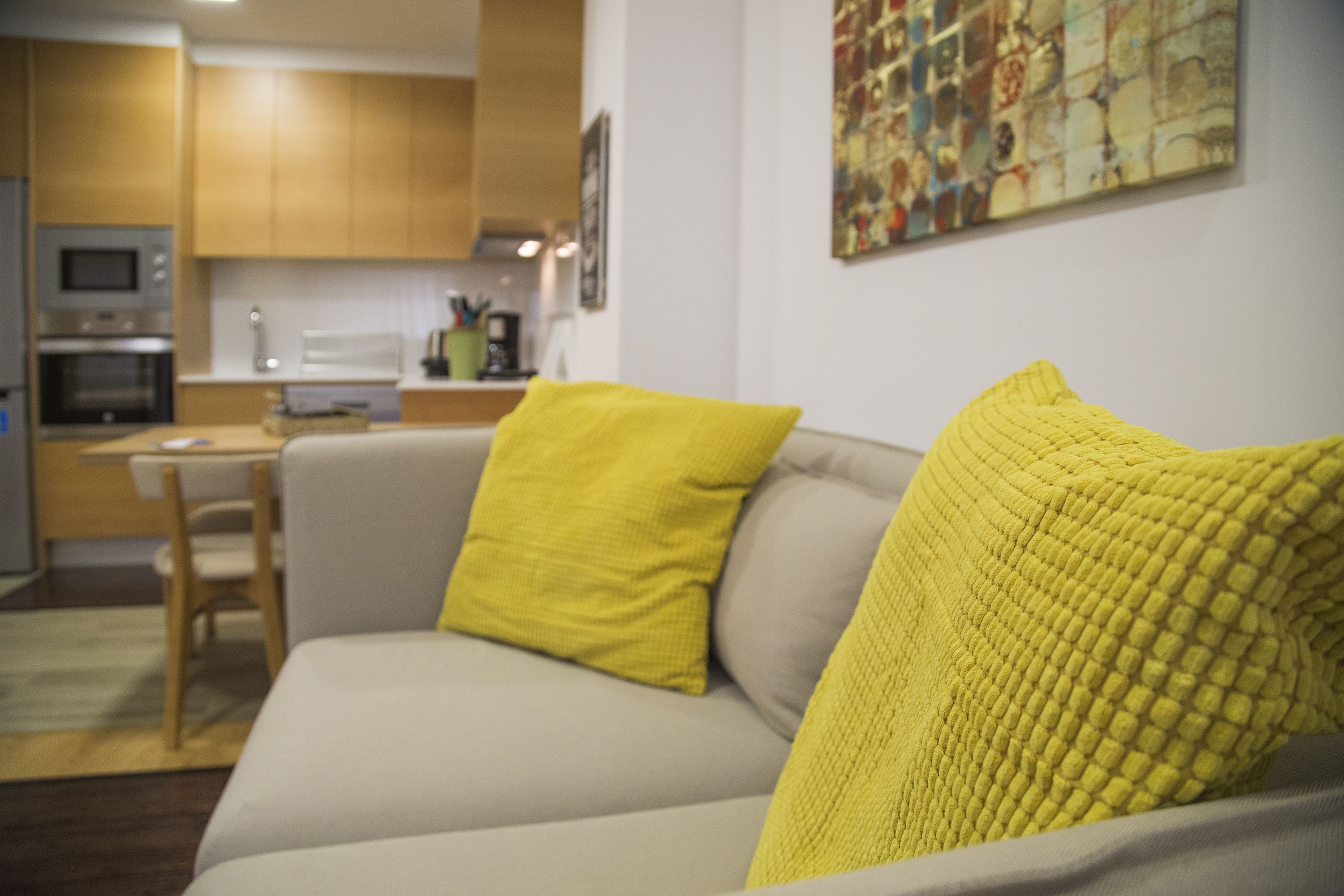 A living room with a sofa, yellow cushions, a kitchen in the background, and a colorful abstract painting on the wall.