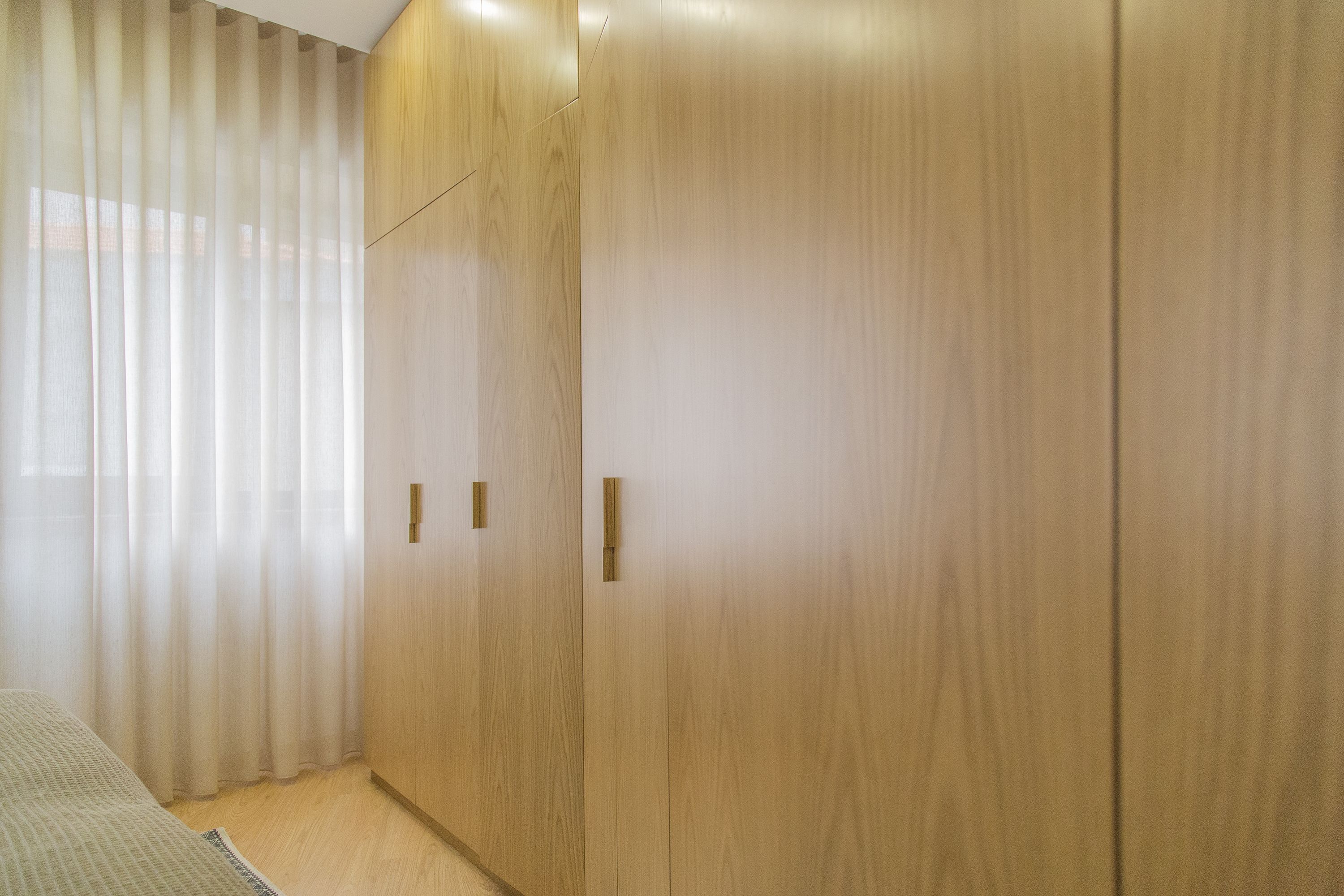 A room with wooden wardrobes, a bed, and sheer curtains.
