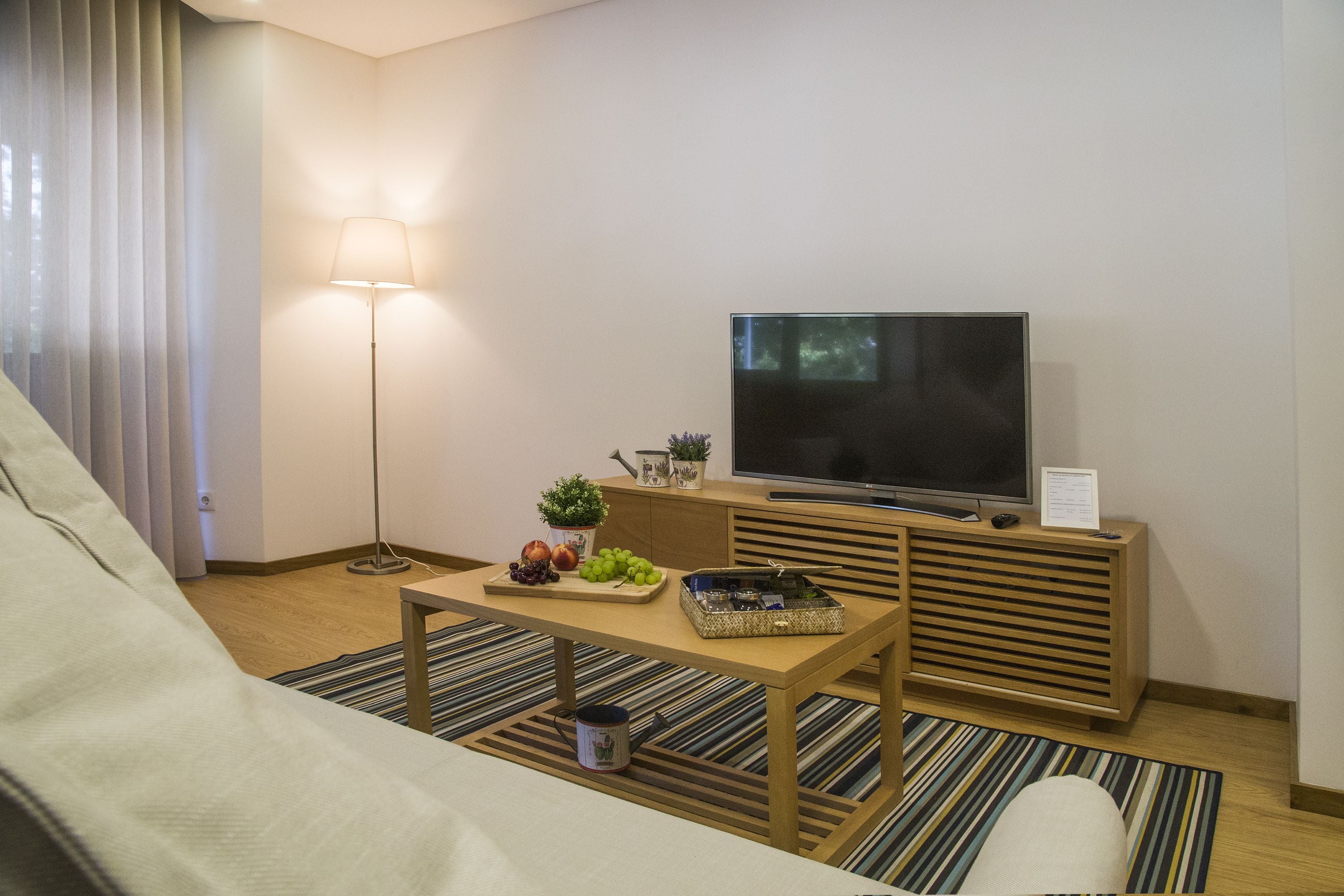A modern hotel room with a flat-screen TV, a wooden coffee table with fruits and a basket, a floor lamp, and a sofa.