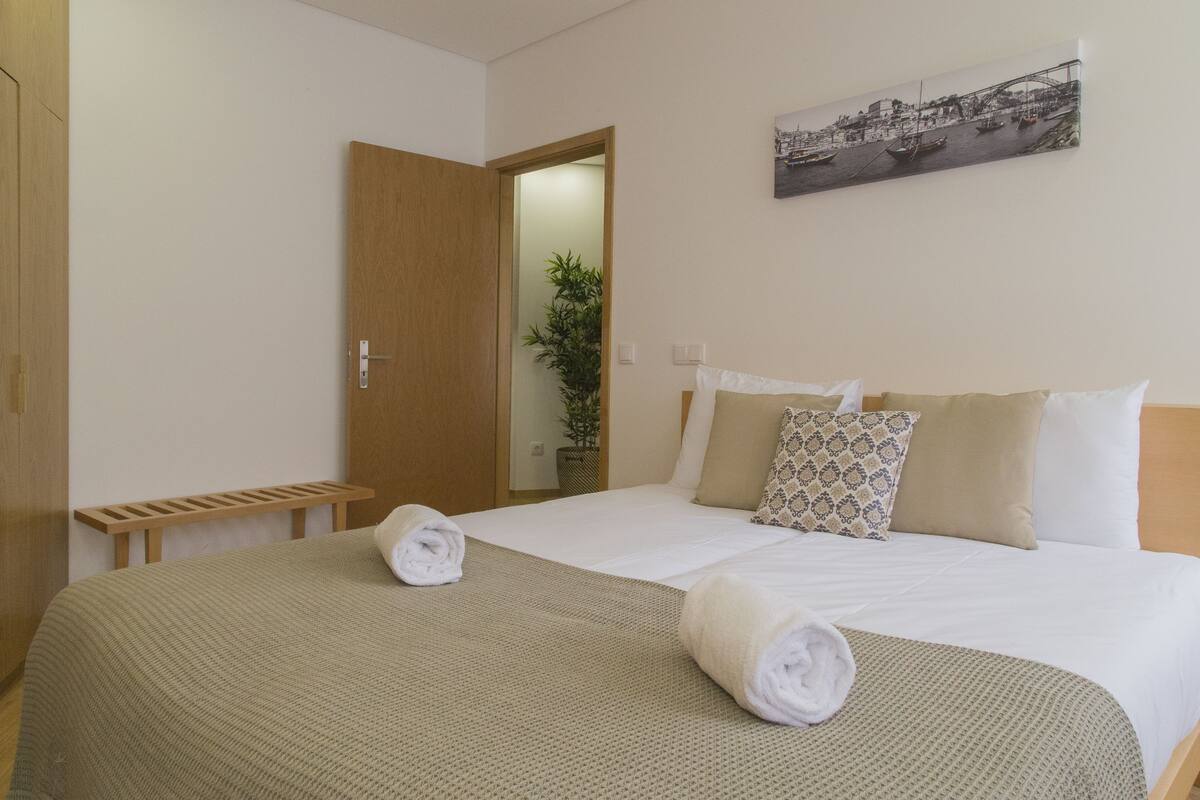 Ein ordentlich bezogenes Bett mit einer beigen Decke und zwei aufgerollten Handtüchern, eine hölzerne Bank und eine Tür zu einem anderen Raum.