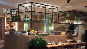 A modern bar area with a wooden counter, glass shelves holding bottles and glasses, and a variety of food items on the counter. - Spot Apartments Trindade (Porto)