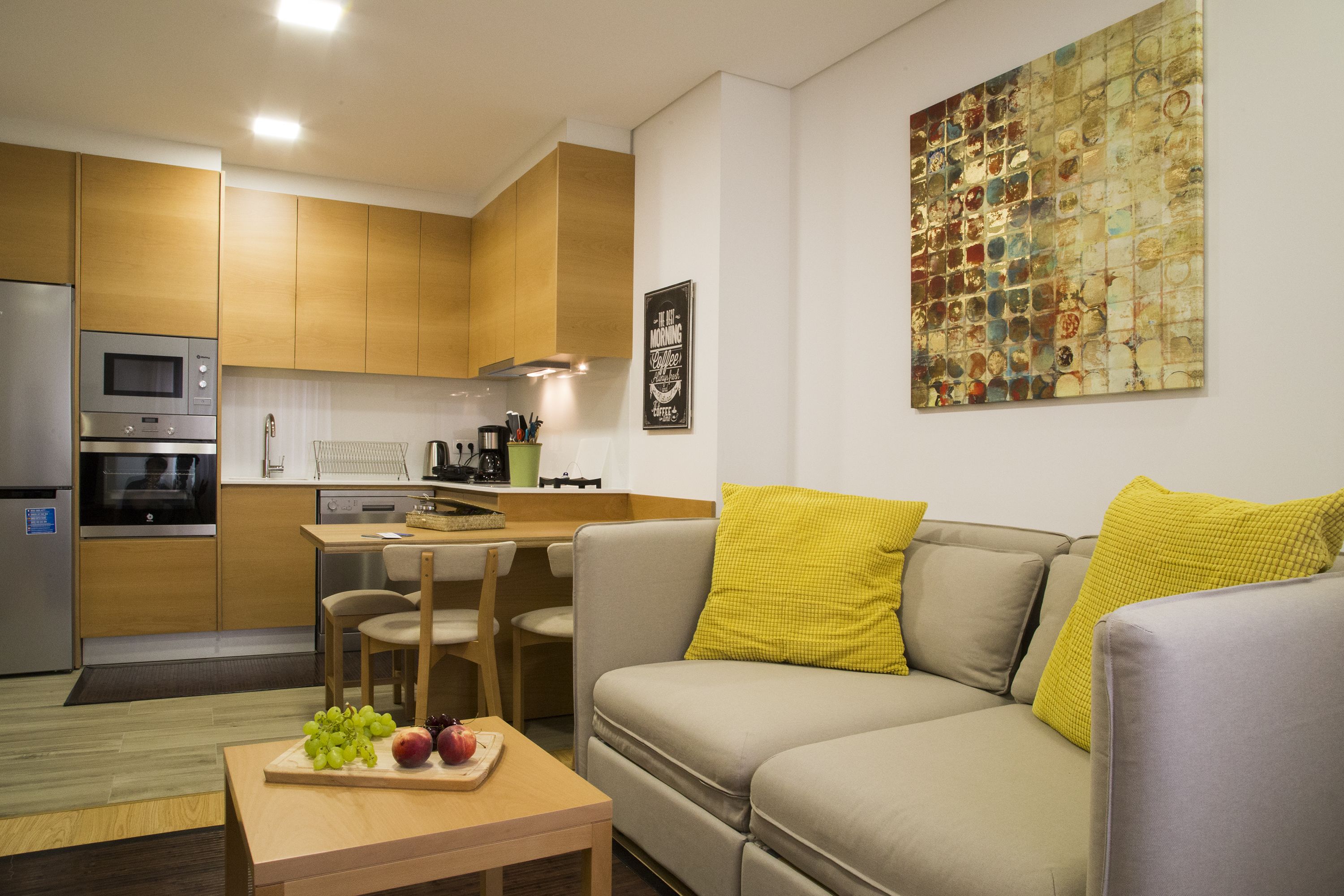 A modern kitchen with wooden cabinets, a dining area with a table and chairs, and a living area with a sofa and cushions.